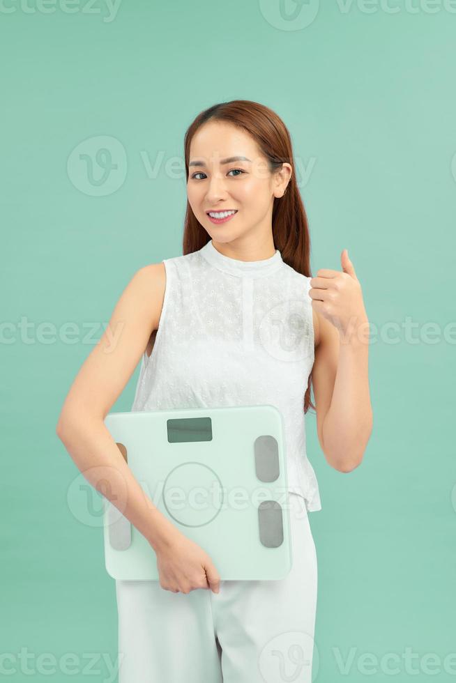 A young happy female holding a weight scale and giving a thumb up isolated on blue background photo