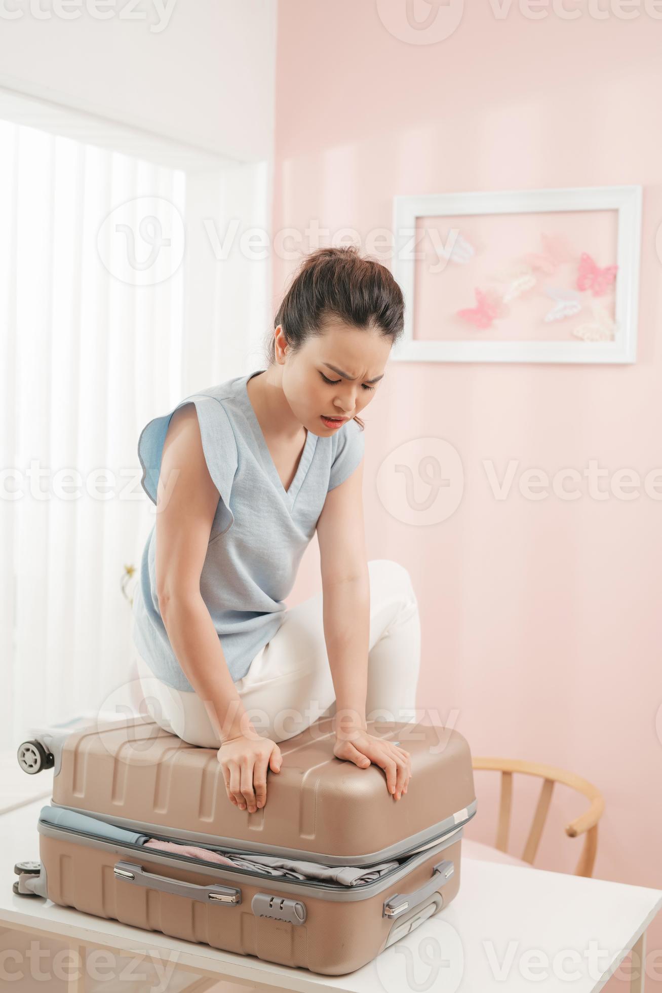 Sad young woman sitting on stuffed suitcase, smiling. Girl packing