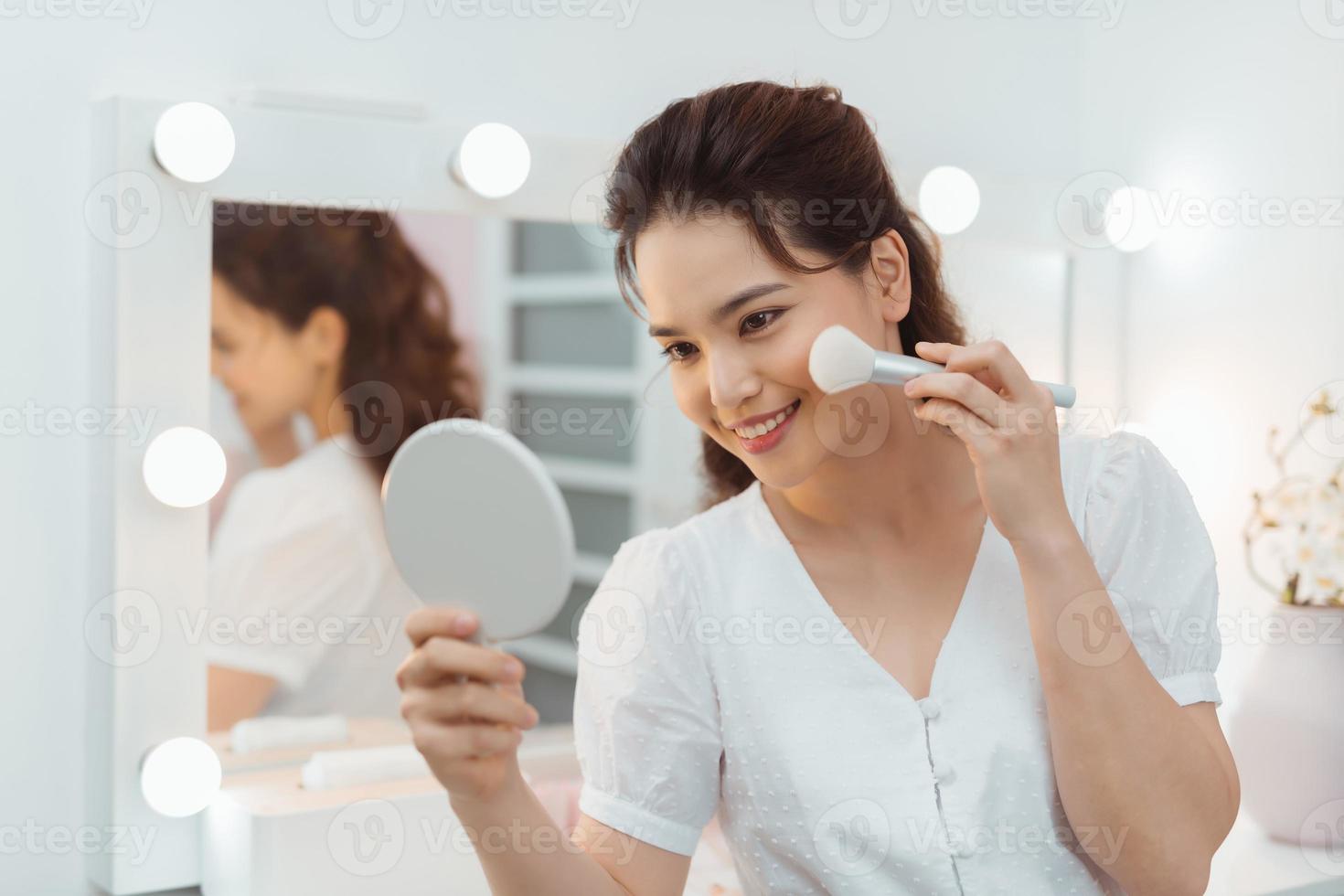 Beauty model girl applying makeup and smiling. Beautiful young woman looking in the mirror and applying cosmetic with a big brush. photo