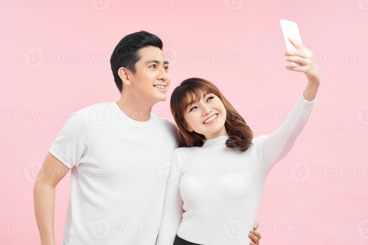 Smiling asian couple taking a selfie isolated over pink background photo