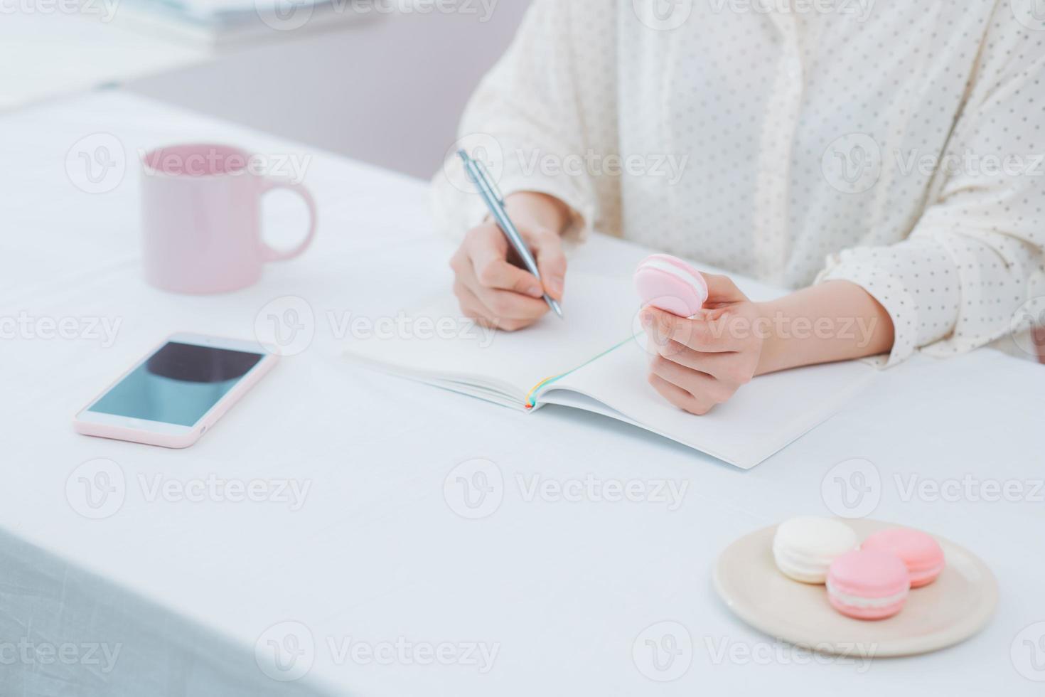 primer plano de mujer o estudiante escribiendo en cuaderno con lápiz, taza, macarons, teléfono foto