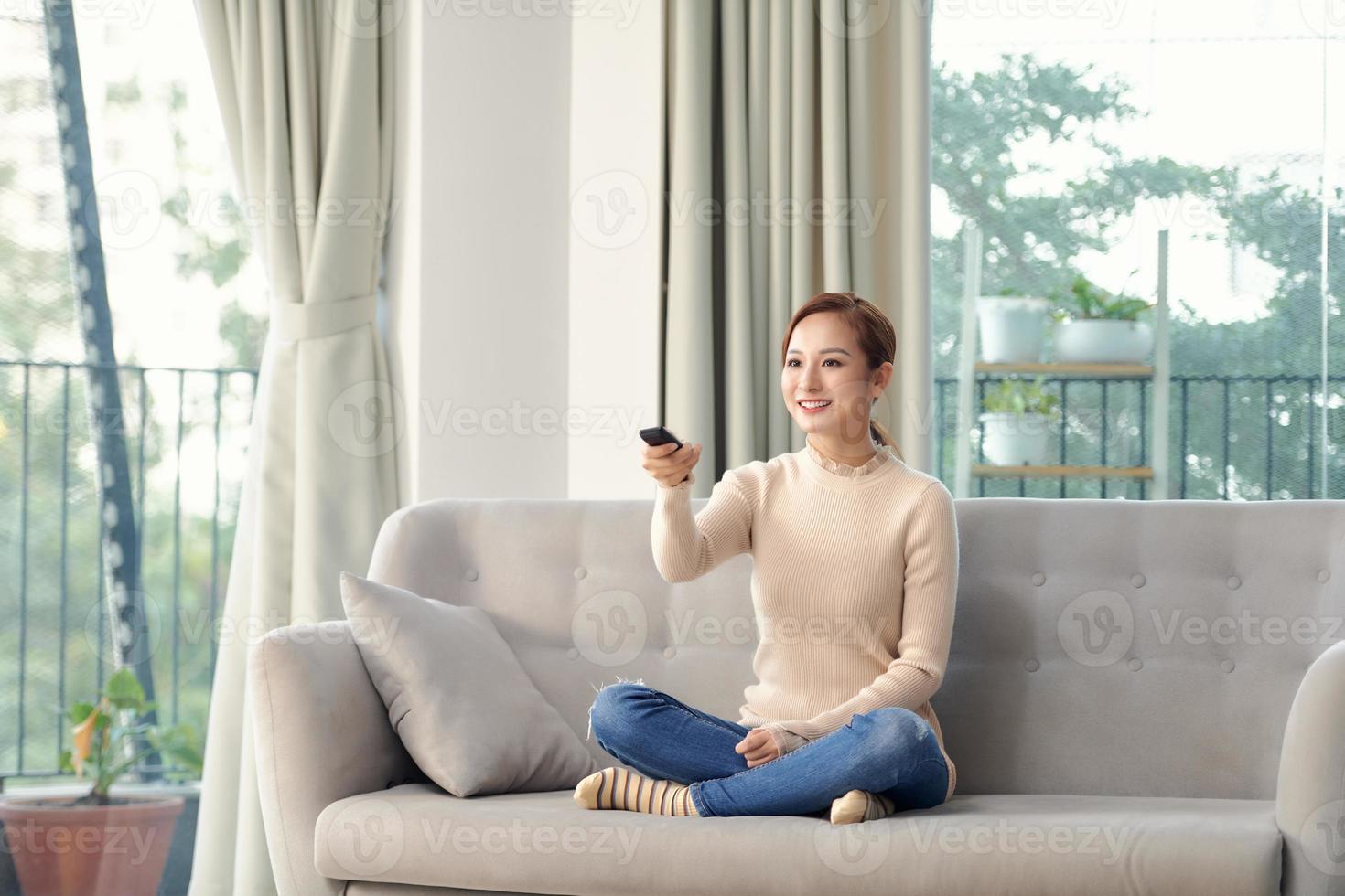 Photo of happy young woman sitting on sofa at home. Looking camera holding remote control watch TV.
