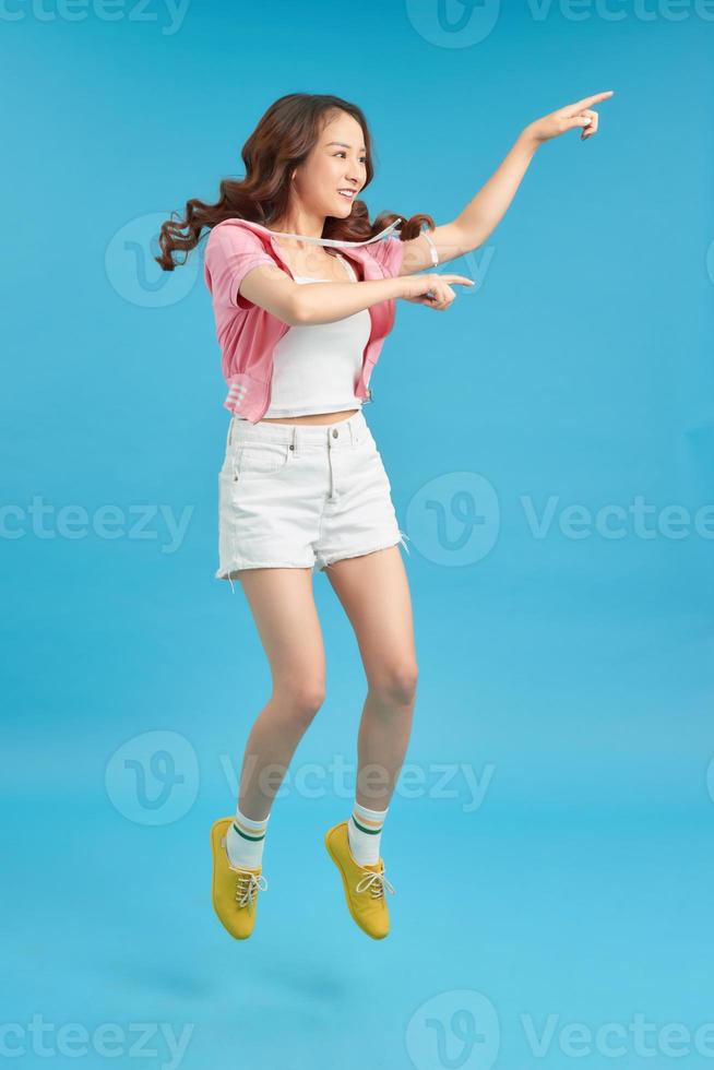 Full length image of Joyful asian woman jumping while pointing and looking away over blue background photo