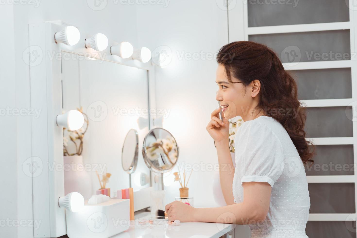 Amazing young woman doing her makeup in front of mirror. Portrait of beautiful girl near cosmetic table photo