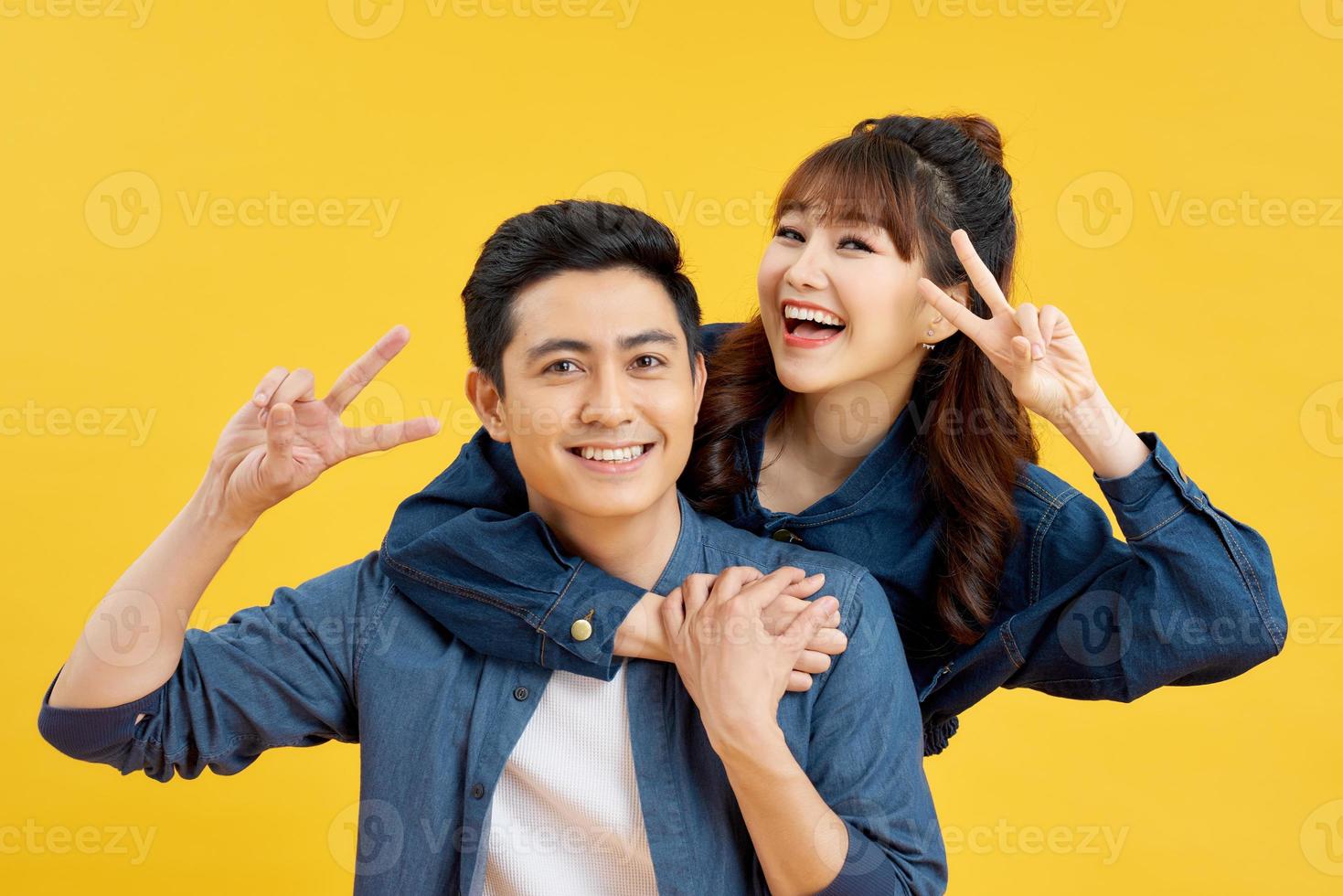 He and she travel together, love story of attractive, happy, smiling, sweet couple in casual outfit, men carrying his lover on back, woman showing peace symbol over yellow background photo