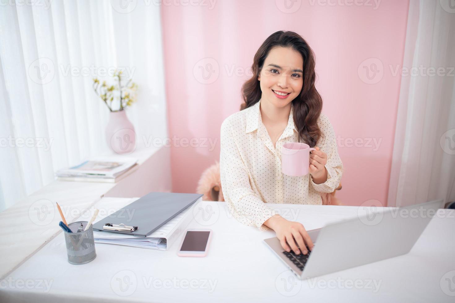 hermosa mujer asiática usando una laptop en su oficina mientras sostiene una taza de té. foto