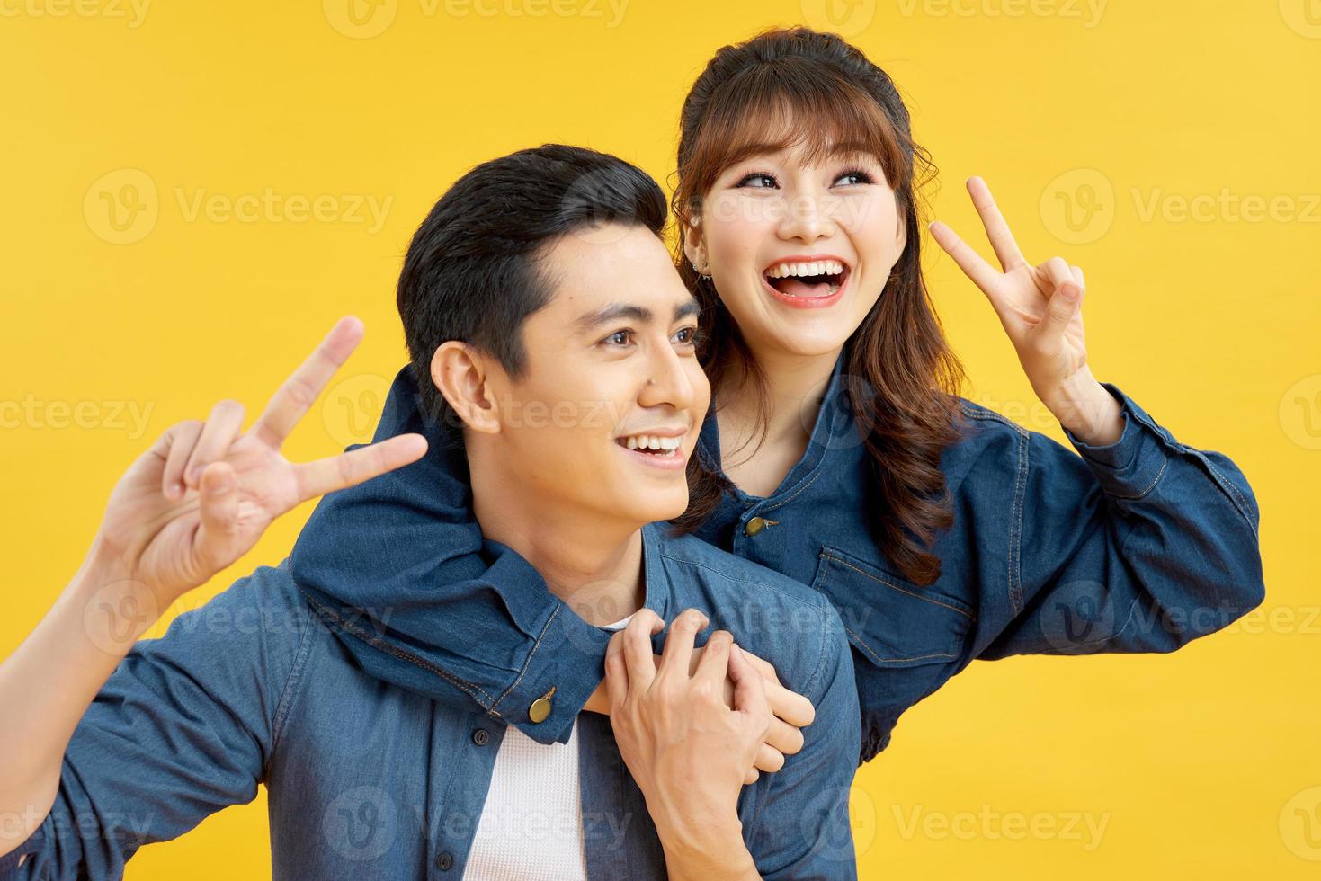 He and she travel together, love story of attractive, happy, smiling, sweet couple in casual outfit, men carrying his lover on back, woman showing peace symbol over yellow background photo