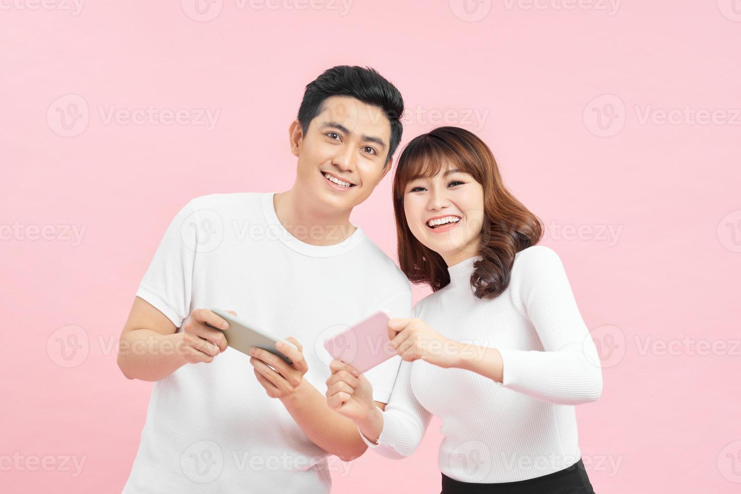 Beautiful couple wearing white t-shirts standing isolated over pink background, playing games on mobile phone. photo