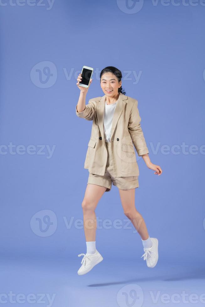 Happy asian young woman showing empty smartphone screen while jumping up over purple background photo