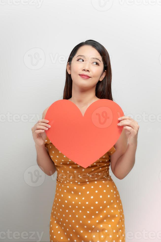 Young asian woman isolated on white wall concept holding heart close-up thinking photo