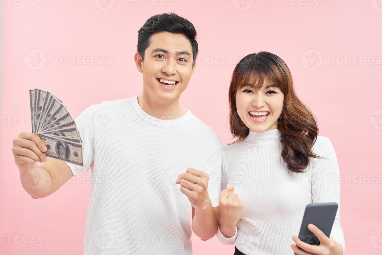 Photo of happy young loving couple standing isolated over pink background holding money showing display of mobile phone pointing looking camera.