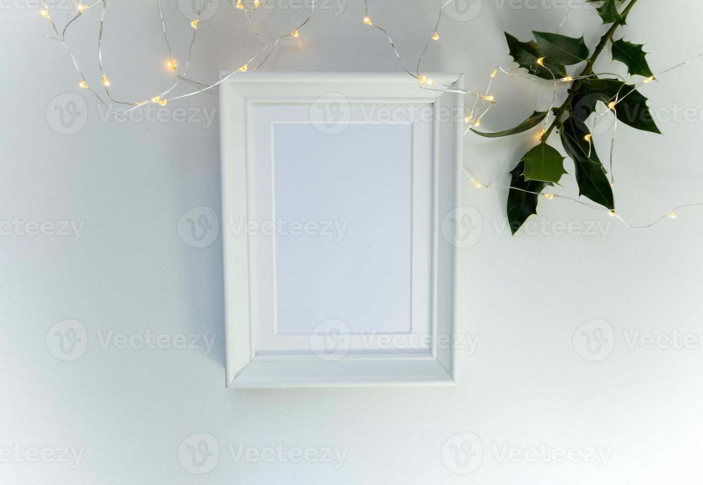 Blank white photo frame with a holly plant branch and lights garland, mockup to display illustrations, art products, simple minimalist concept