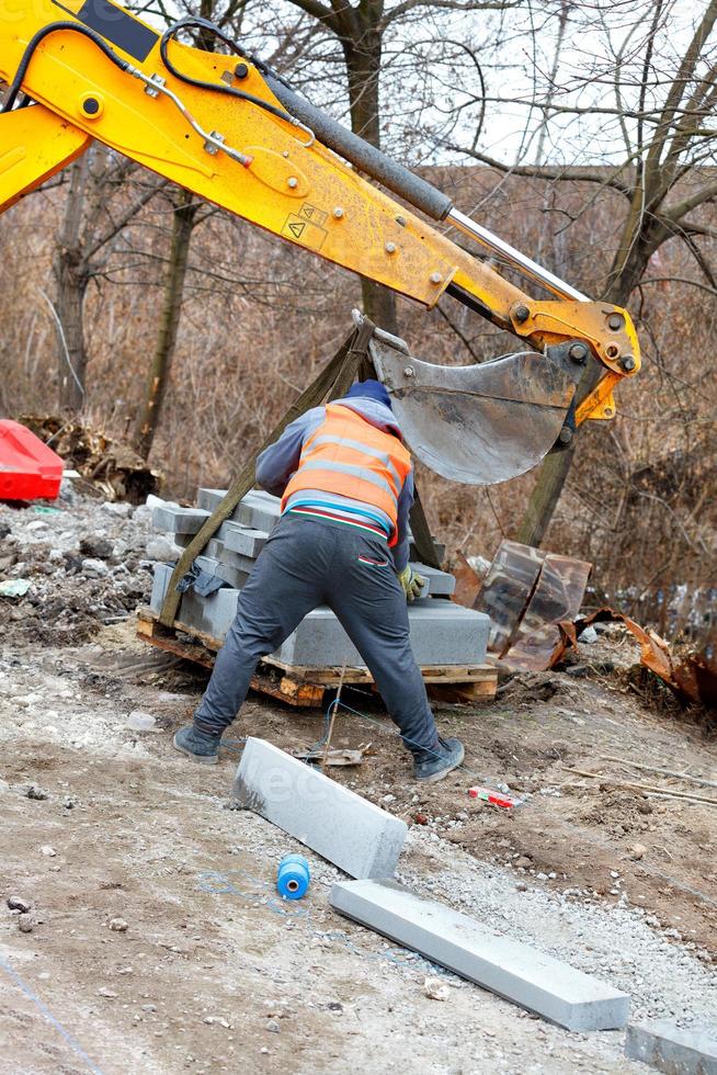 A construction worker using an excavator boom and bucket unloads concrete curbs at a work site. photo