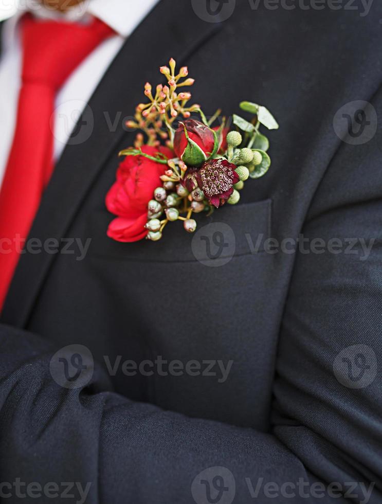 The groom's wedding boutonniere of succulents and red flowers in a black jacket with a red tie. Festive decor, flowers, attire for the marriage registration. Close-up, space for text photo