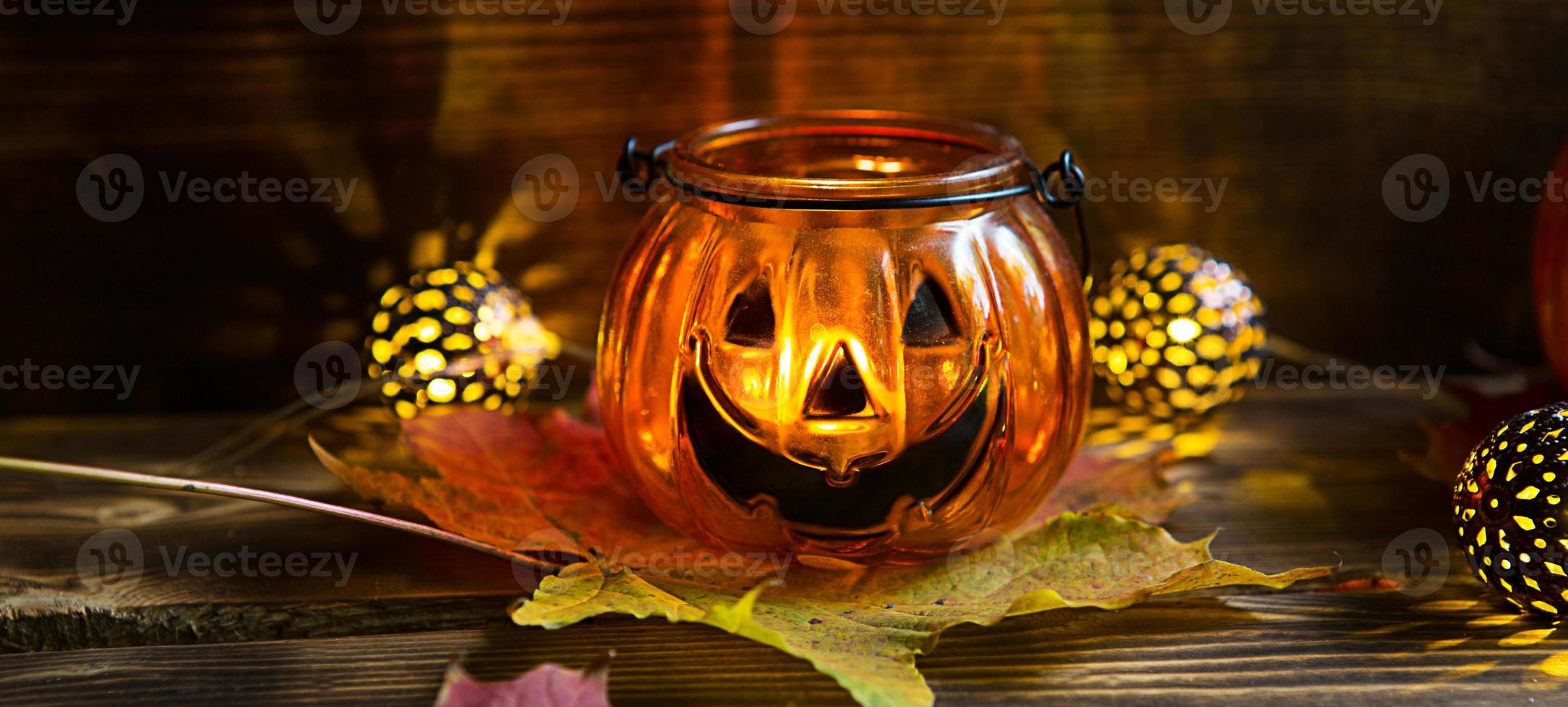 Lamp pumpkin with eyes and mouth made of glass and natural orange pumpkin on a wooden table with yellow and red maple leaves. Halloween, warm autumn atmosphere. Round garland, candle in a candlestick. photo