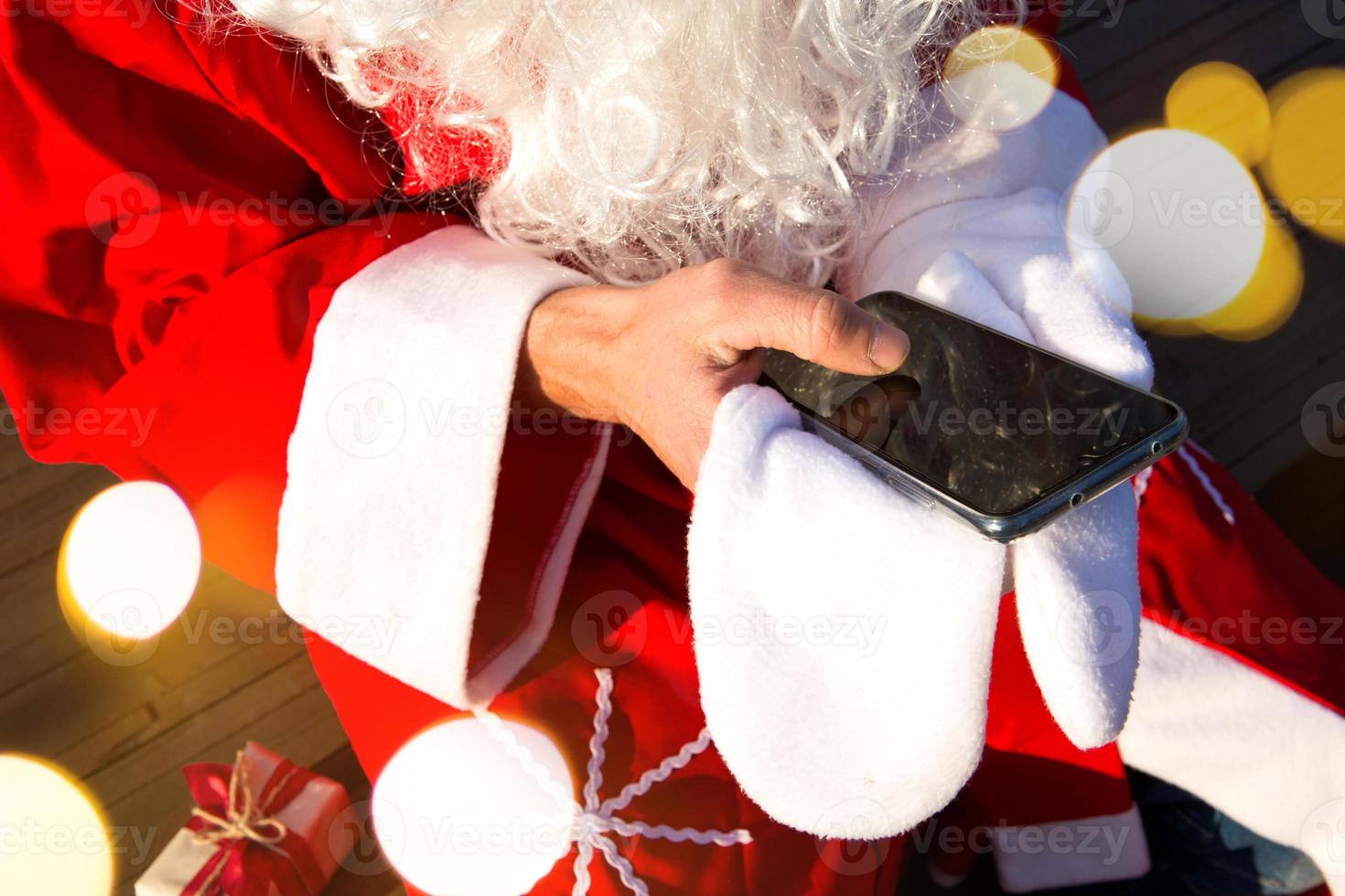 The hand of Santa Claus without a mitten moves his finger across the smartphone screen. Long white beard, red suit. Modern Russian grandfather Frost. New year's order processing photo
