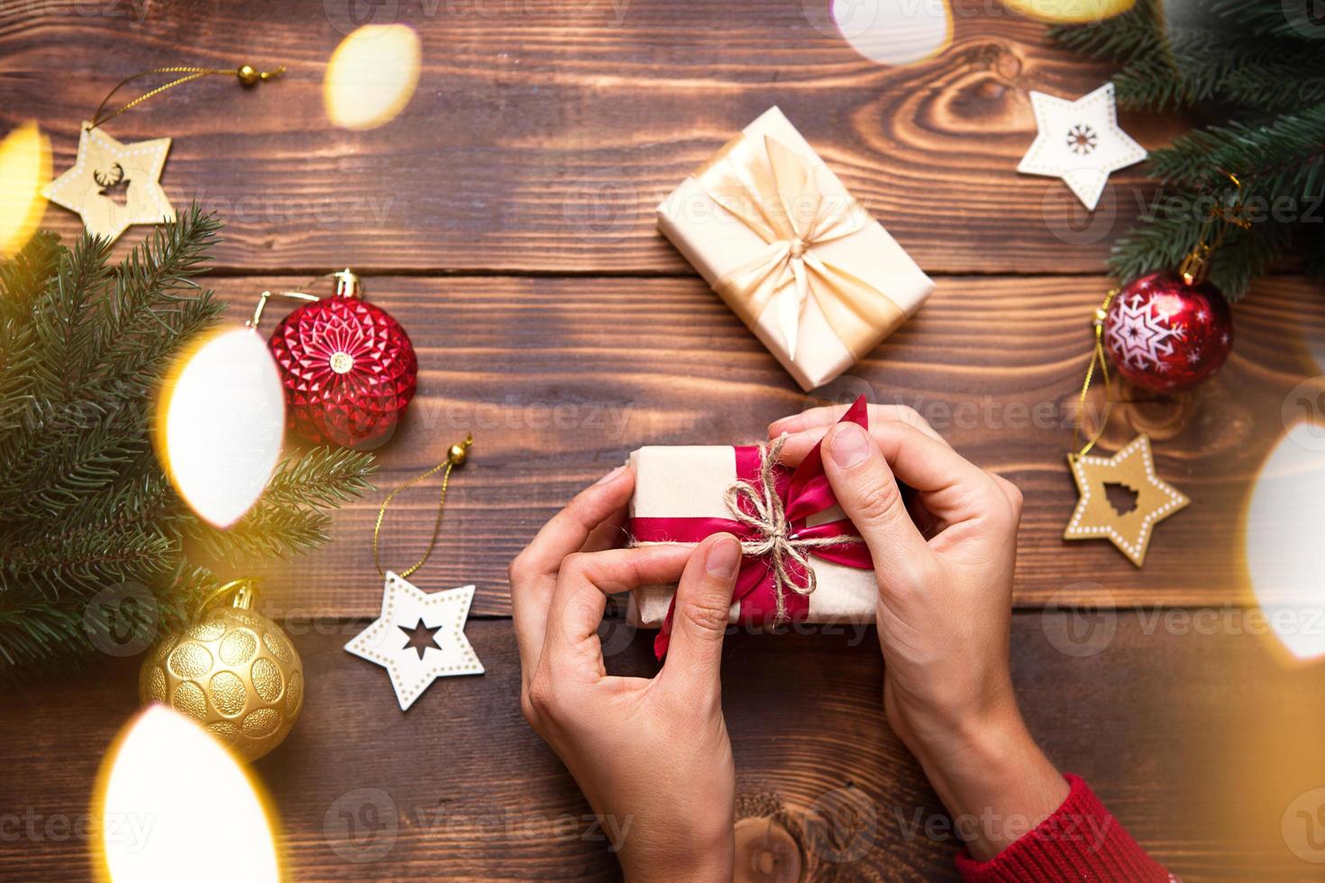 Women's hands in knitted sweater with ornament hold a box with Christmas gift in their palms on the table among the decor. New year, packaging and preparation of gifts, branches of fir, decoration photo
