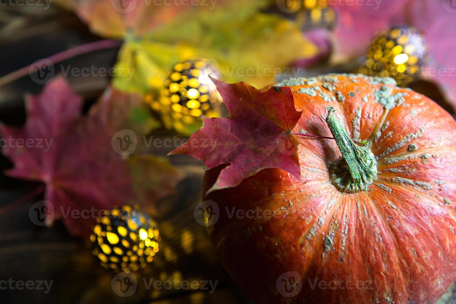 Orange natural round pumpkin on a wooden table with fallen yellow and red maple leaves, cinnamon sticks. Lights garlands, warm autumn atmosphere, thanksgiving, harvest festival, Halloween. Copy space photo