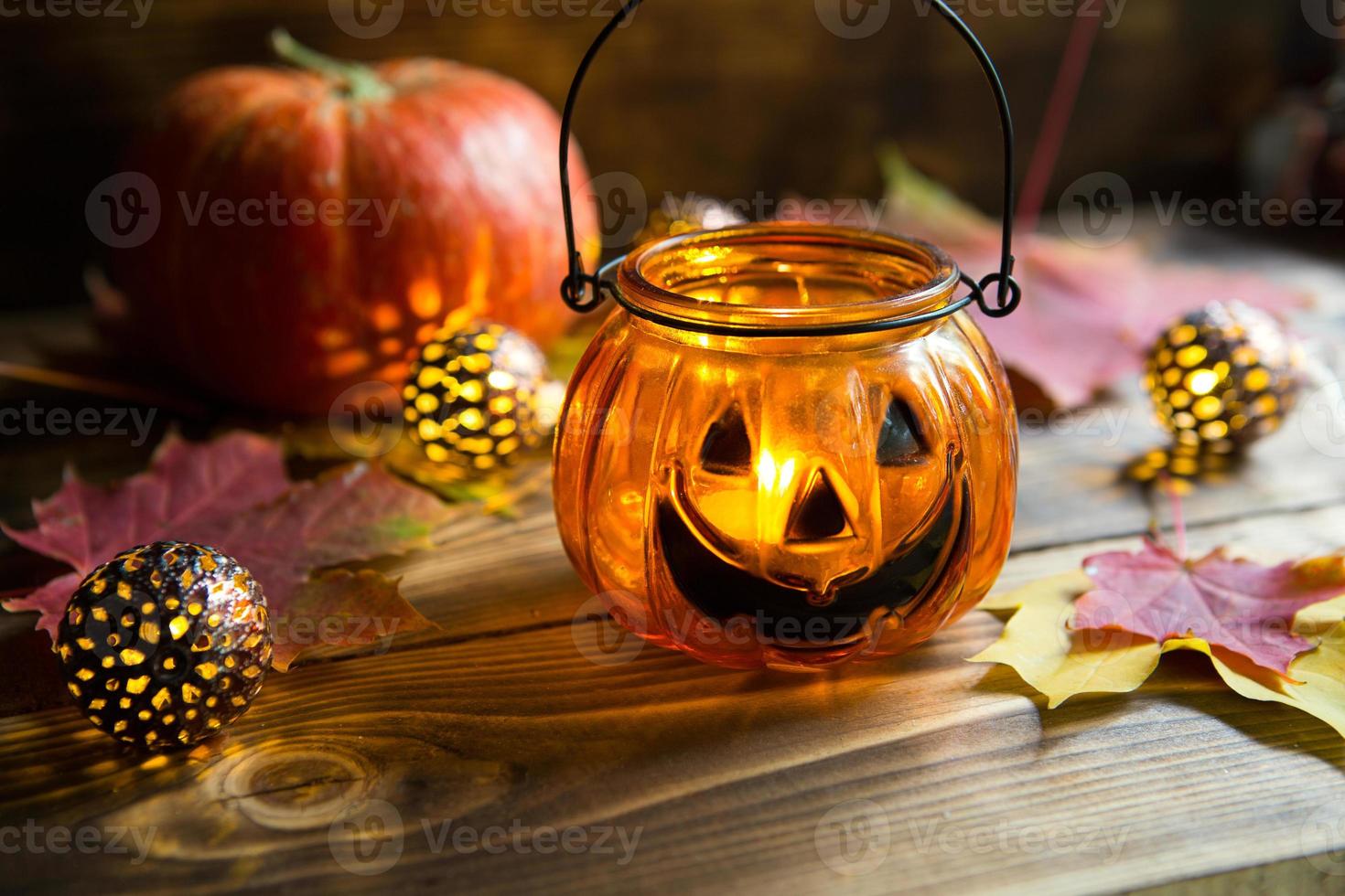 Lamp pumpkin with eyes and mouth made of glass and natural orange pumpkin on a wooden table with yellow and red maple leaves. Halloween, warm autumn atmosphere. Round garland, candle in a candlestick. photo
