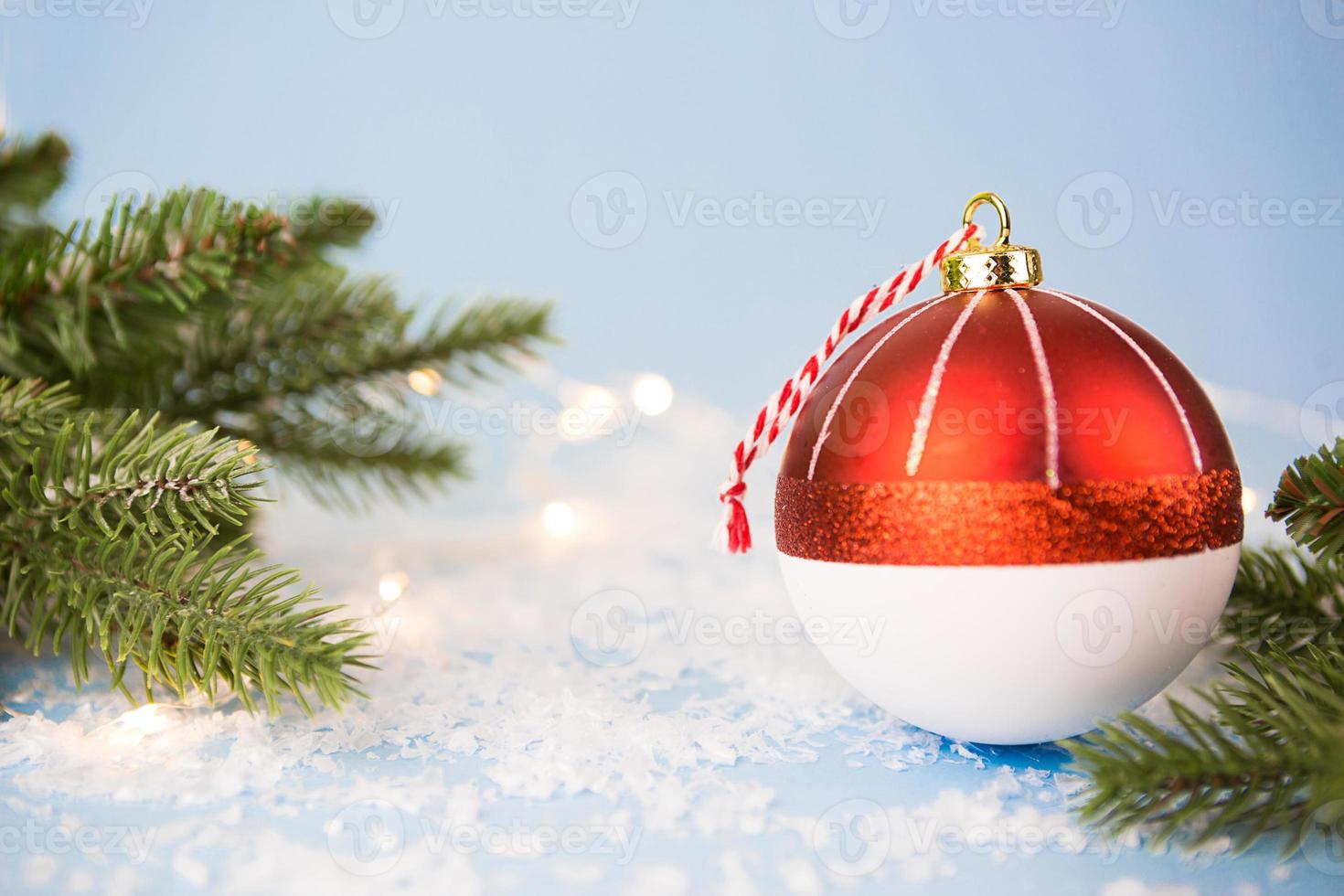 Red Christmas ball on a blue background with artificial snow, the branches of spruce and lights of garlands in bokeh. Festive background, new year. Copy space photo