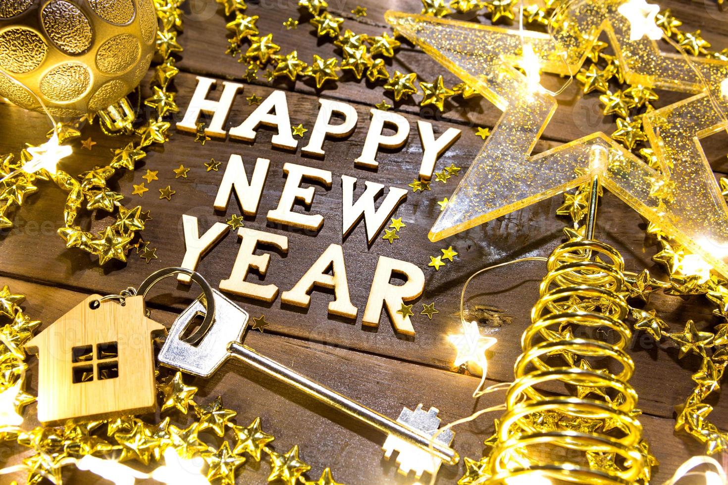 House key with keychain cottage on a festive background with sequins, stars, lights of garlands. Happy New Year-wooden letters, greetings, greeting card. Purchase, construction, relocation, mortgage photo