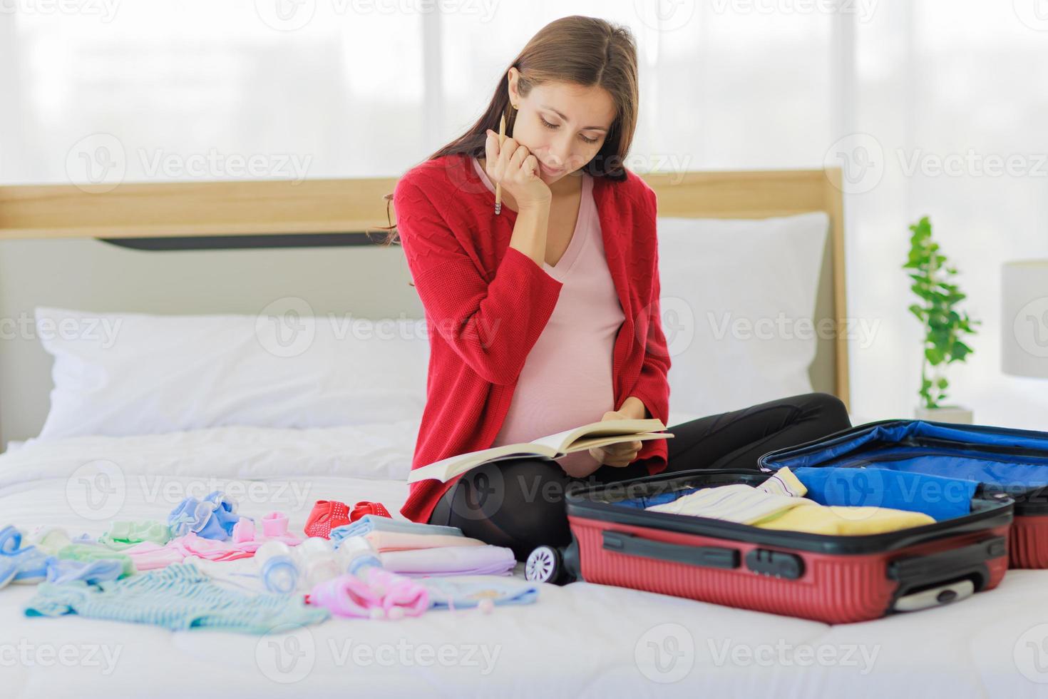 Big pregnant woman happily writing down list of things to prepare baby clothes on her tummy in notebook. photo