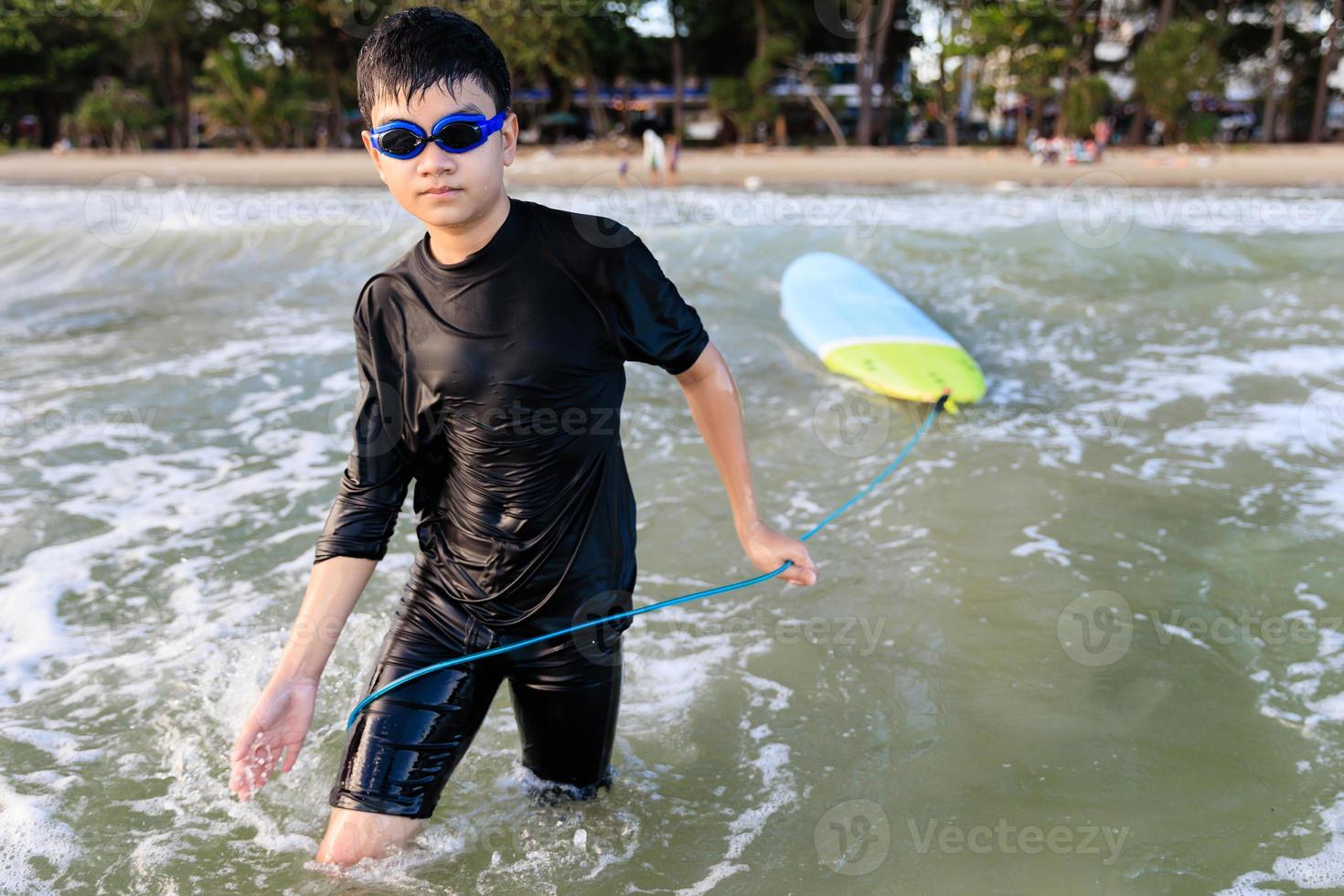 Young teen boy surfer holding rope of soft board, bring it to try again in wave. Rookie teenager surfboard student playing on water in strive focus action. photo
