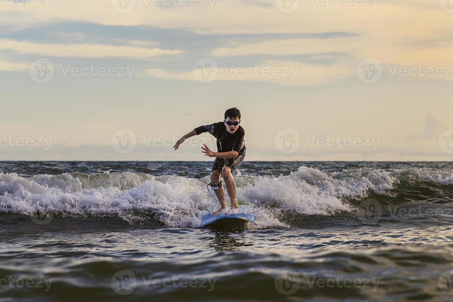 Young boy kid surfer riding waves with a soft board in Rayong beach, Thailand. Rookie surfboard student playing on water in excited face and funny action. photo