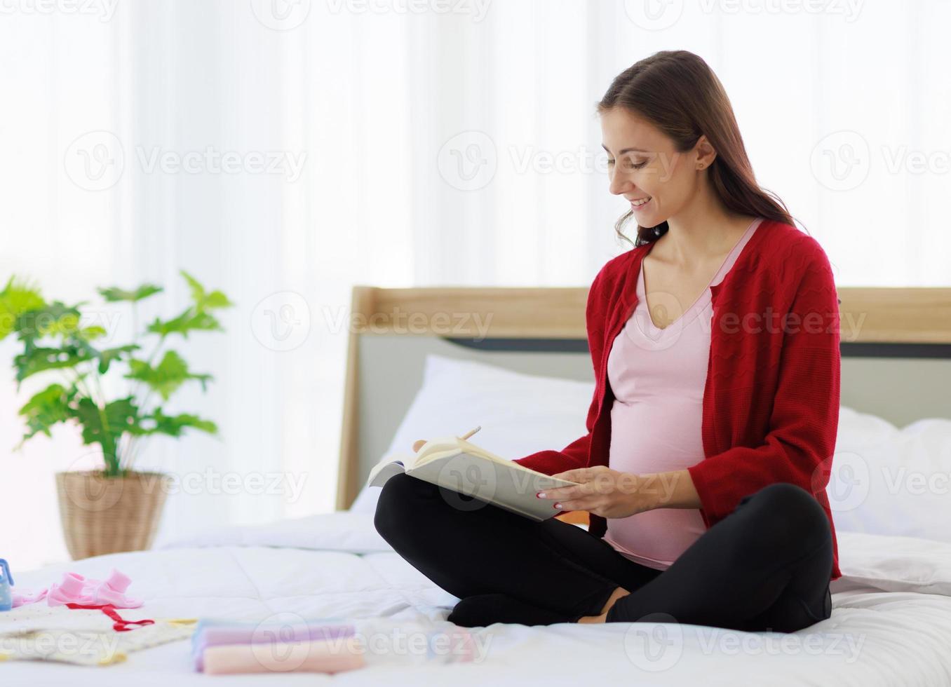 A beautiful, happy caucasian mother is sitting on her bed holding a notebook, keeping a list of all the supplies and clothes in preparation for the birth of new born baby. photo