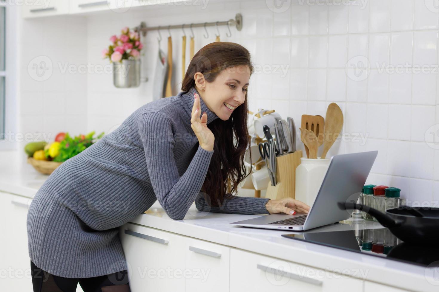 Beautiful, cool, pregnant woman standing in modern kitchen and using laptop computer to chat in videocall with happy and relaxing manner photo