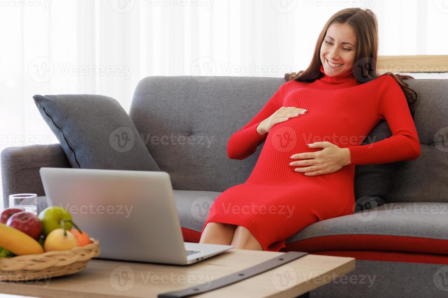 A beautiful, big pregnant woman sits on the sofa and uses her laptop computer to chat on a video call happily and comfortably. Family relationship and emotion care concept photo