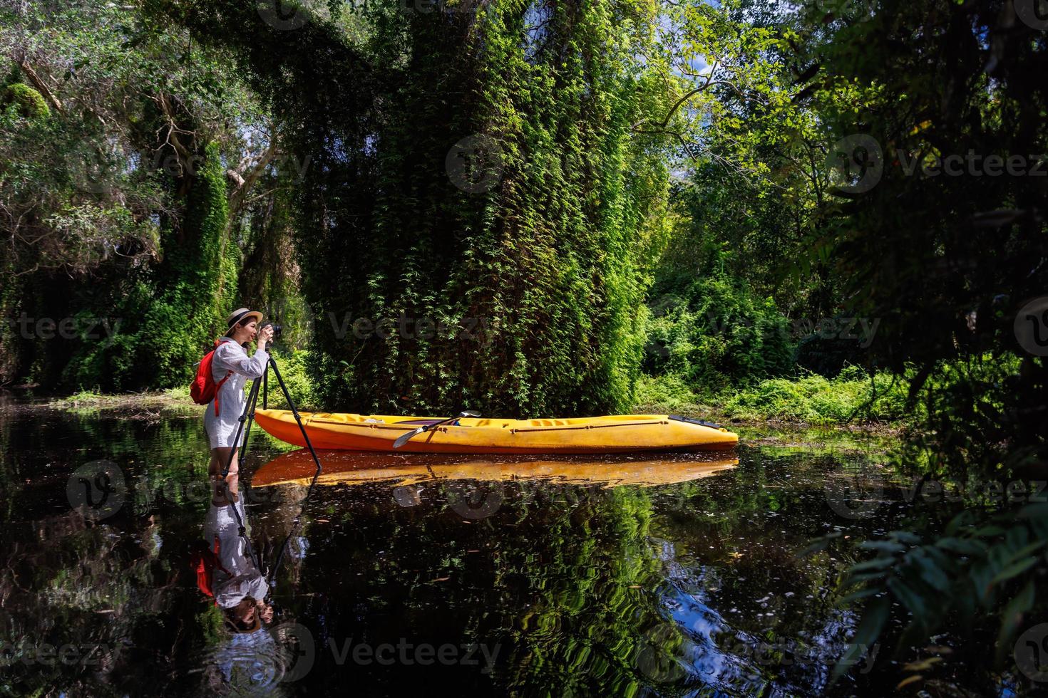 Woman researcher or botanist uses a camera on a tripod to photograph vegetation in a wetland forest. Female tourist stands in a swamp and takes pictures beside a canoe. photo