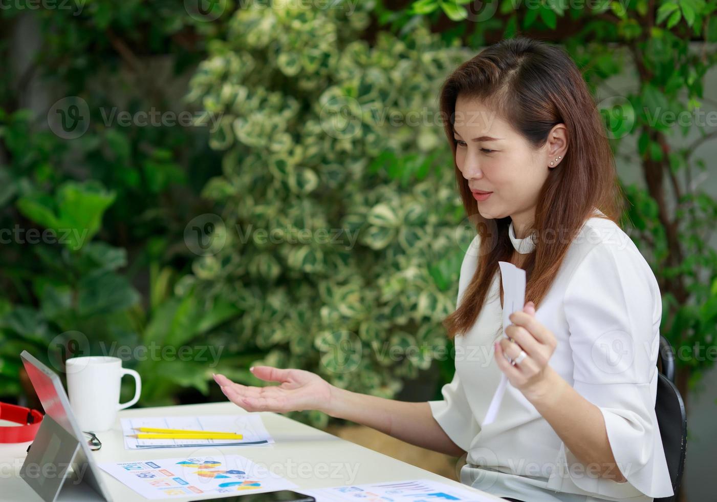 Beautiful smiling Asian businesswoman sitting relaxed in home garden, using tablet and communicate on web call surrounded by exotic plants. photo
