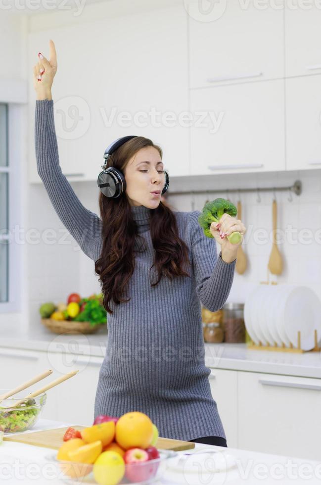 Beautiful, cool, pregnant woman happily cooking organic and healthy salads in her modern kitchen. New mom uses broccoli instead of microphone to sing and play for fun. Good mood concept photo