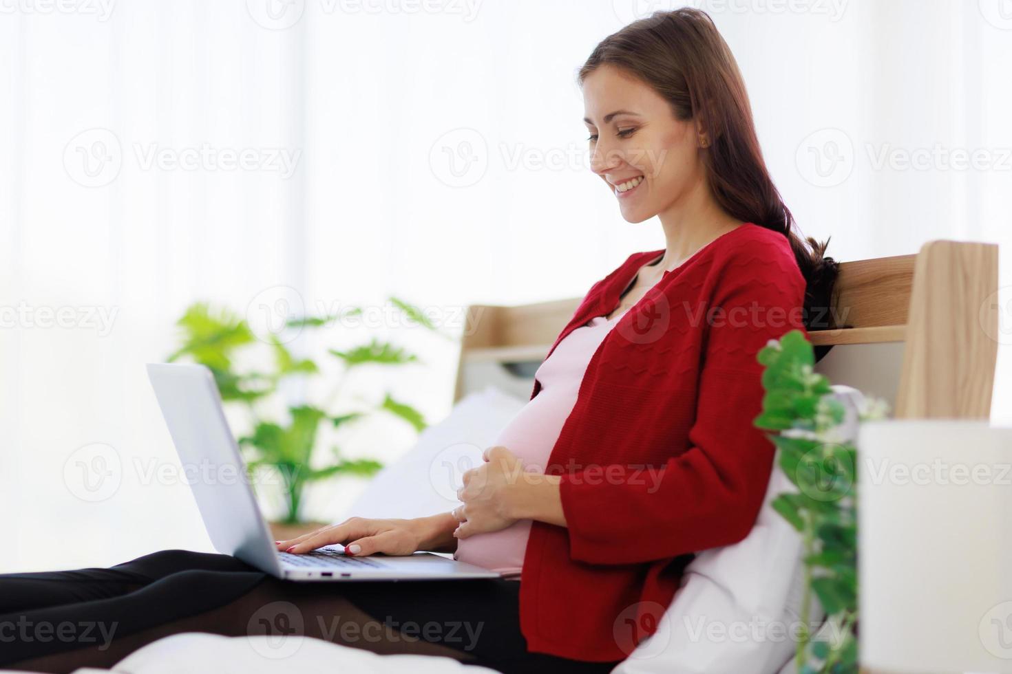 una hermosa y feliz madre caucásica se sienta en la cama usando su computadora portátil para buscar información. foto