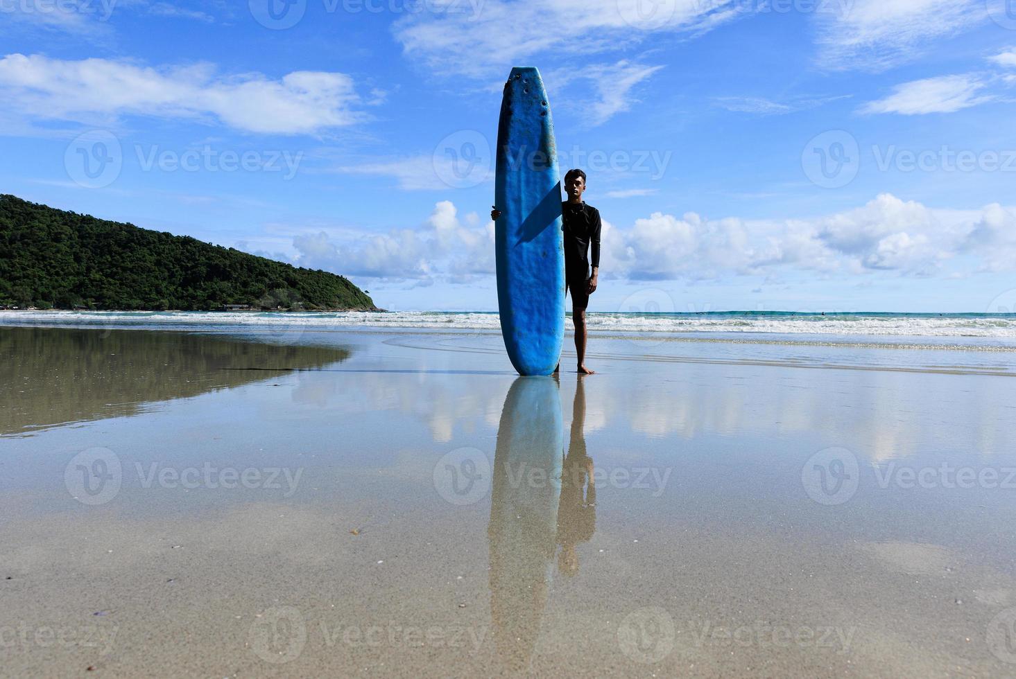 A young Asian teenage guy standing with self-confidence holding a soft board ready for extreme sports surfing on beach holidays. photo