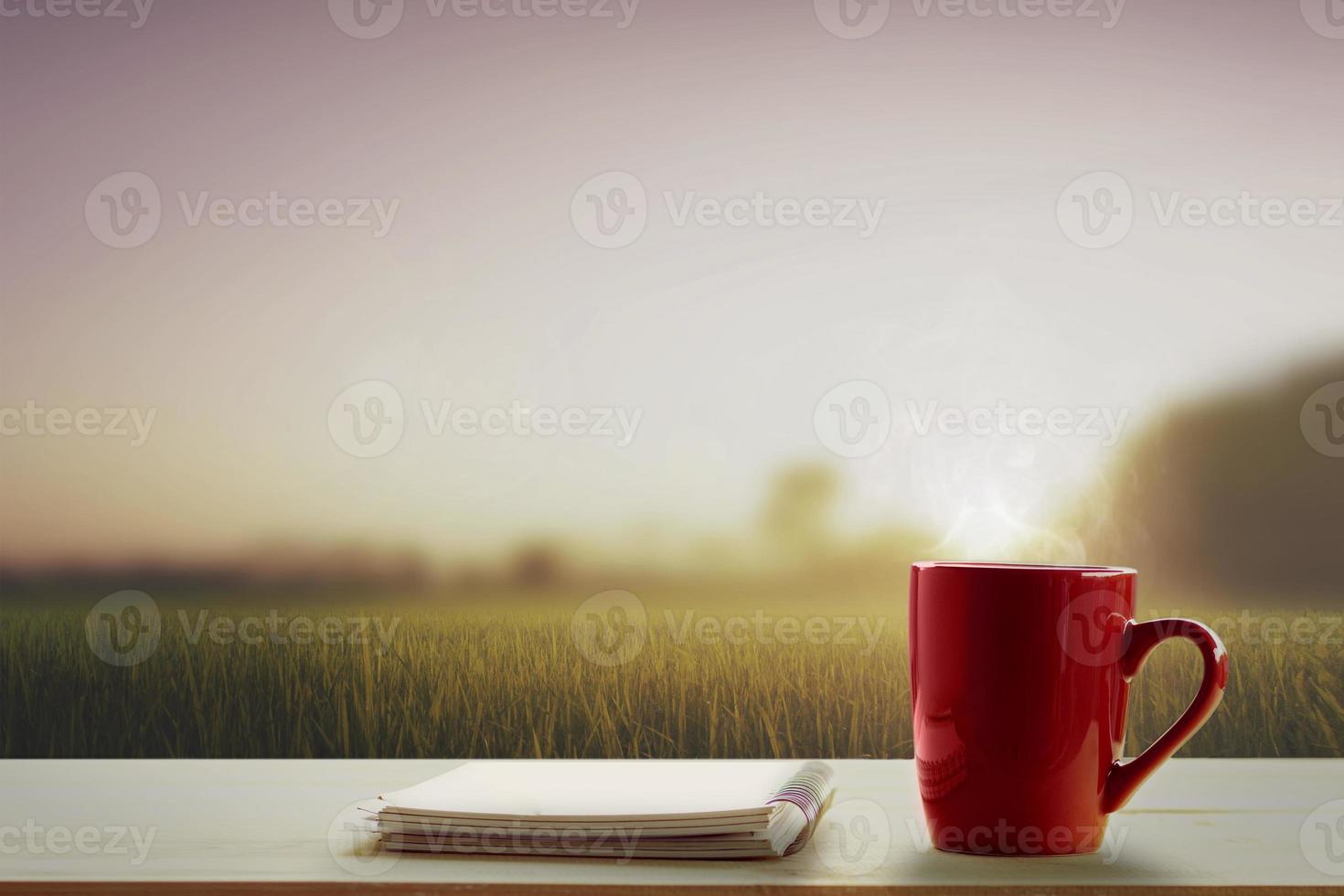 a red cup of coffee and note pad on wooden table and meadows at morning time blurry background photo