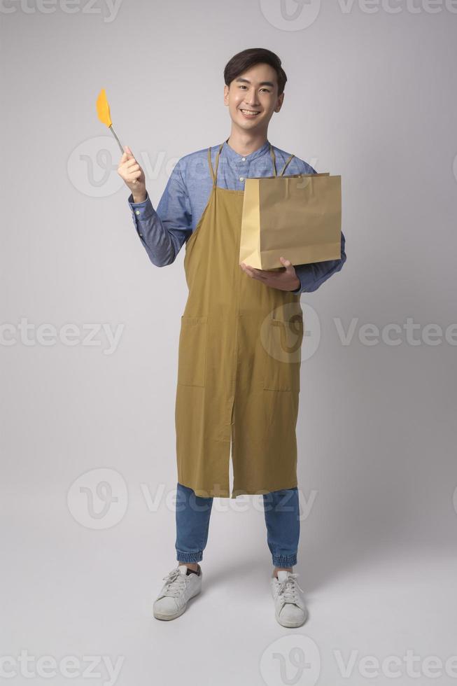 Portrait of young asian man wearing apron holding  paper bag and turner over white background studio. photo