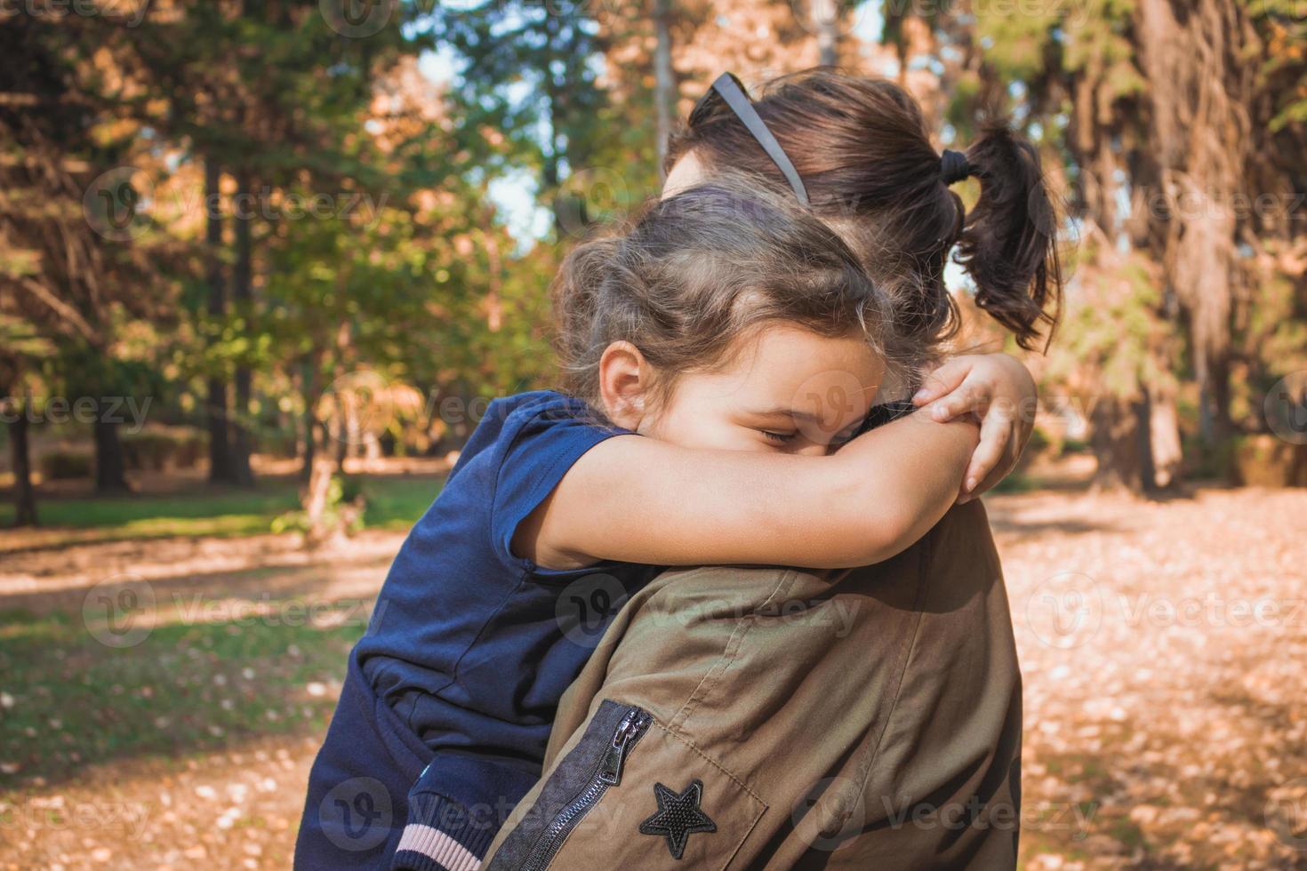 Displeased small girl embracing mother with her eyes closed. photo