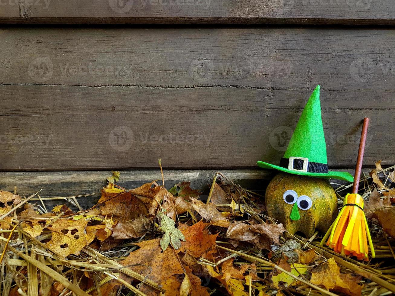 Green funny Halloween pumpkin with goggle eyes in green witch felt hat, her yellow broomstick is near. Old dark wooden wall background with copy space. photo