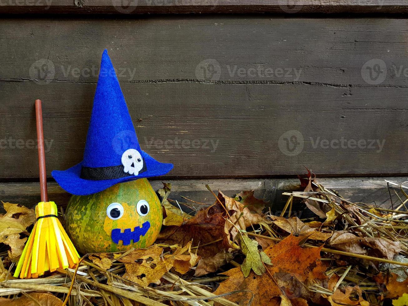 Funny Halloween green pumpkin with goggle eyes in blue witch felt hat and skull on it, yellow broomstick near. Old dark wooden wall background with copy space. Withered foliage and straw underneath. photo