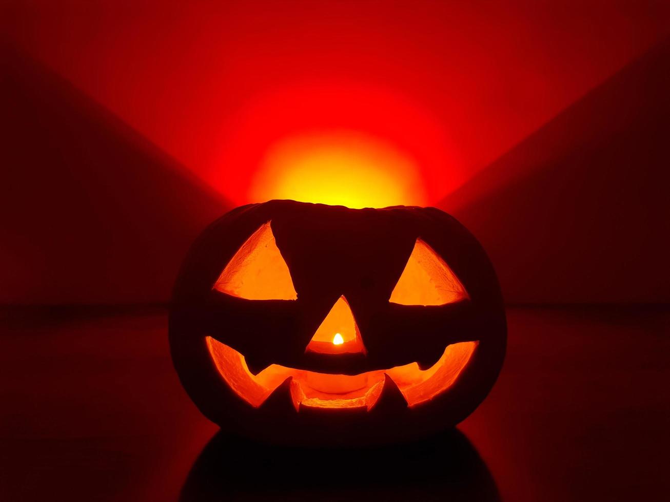 Carved glowing pumpkin with candle inside in dark night and blurred defocused background. The back wall over pumpkin highlighted in orange red gradient triangle. Halloween traditional jack-o-lantern. photo