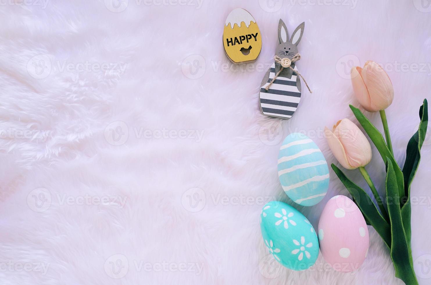 Happy Easter concept with wooden bunny and colorful easter eggs on white fur background and pink tulips. Top view with copy space photo