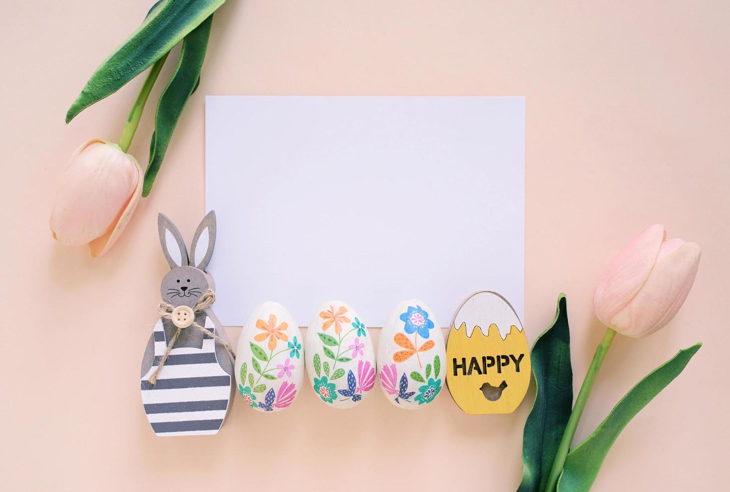 feliz concepto de pascua con tarjeta en blanco, conejito de madera, coloridos huevos de pascua y tulipanes rosas. vista superior con espacio de copia foto