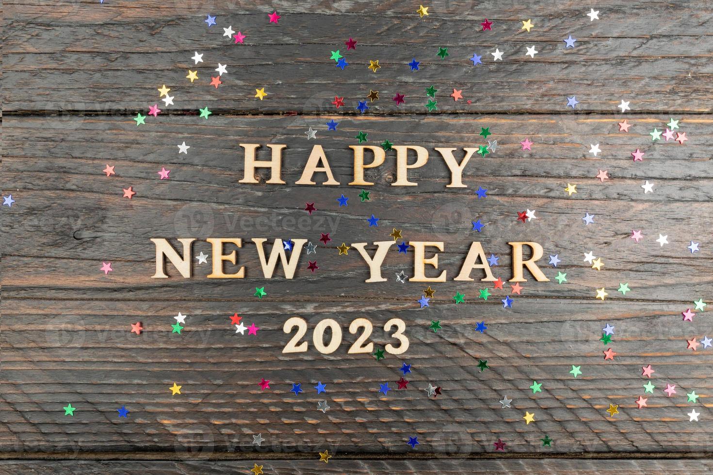 Happy new year 2023 quotation on wooden background with scattered confetti of multicolored stars. Festive greeting card photo