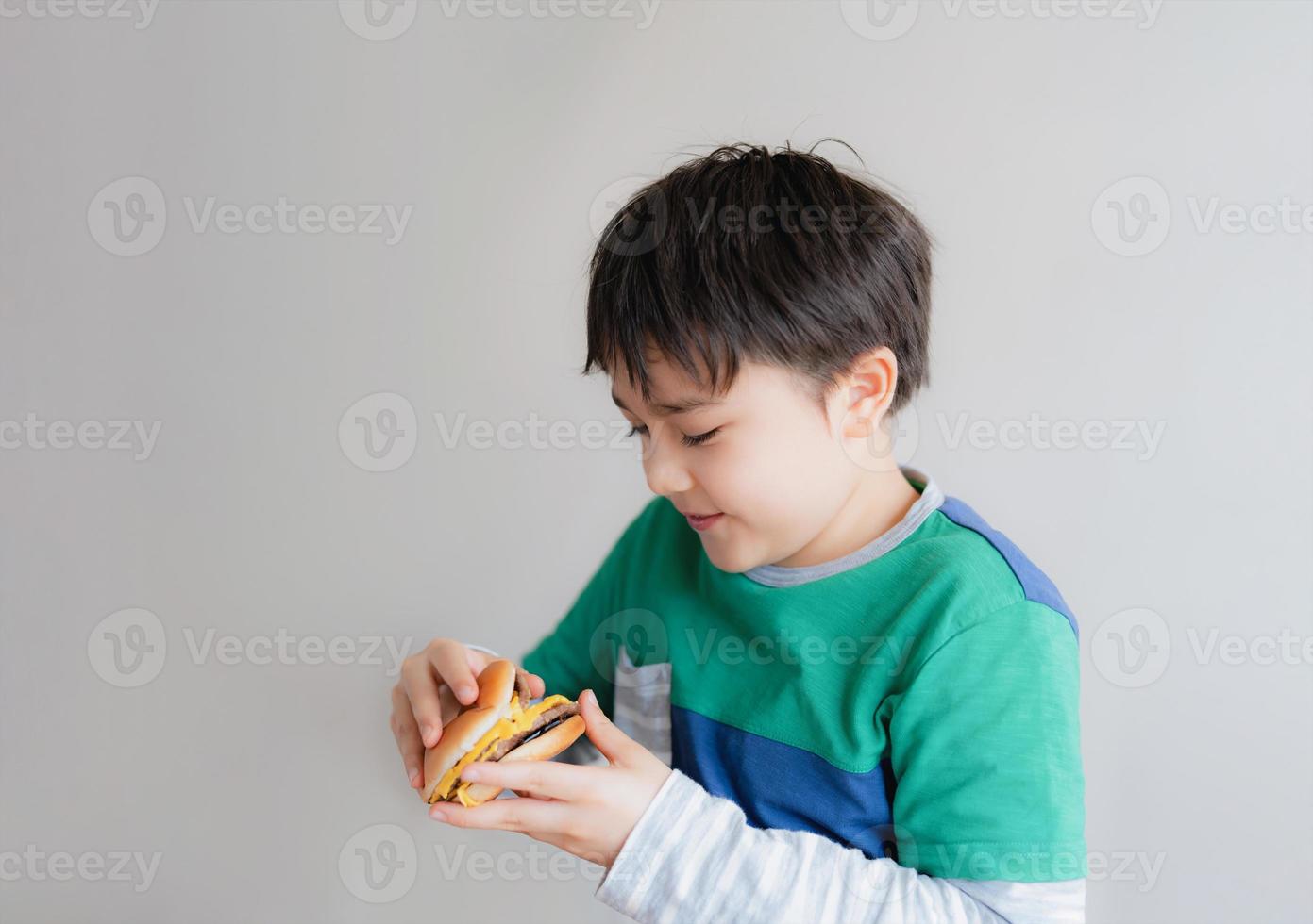 Healthy young boy eating burger. A child holding cheeseburger and looking with smiling face, Isolated Happy Kid having fast food hamburger for snack. Favorite children's food. photo