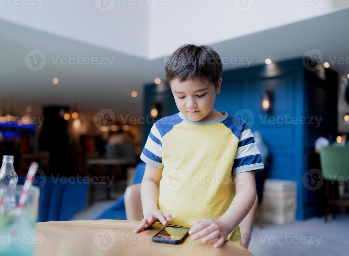 Portrait young boy playing game on mobile while waiting for food, Kid sitting in coffee shop sending text to friends, Child playing game online on cellphone.Children with technology concept photo