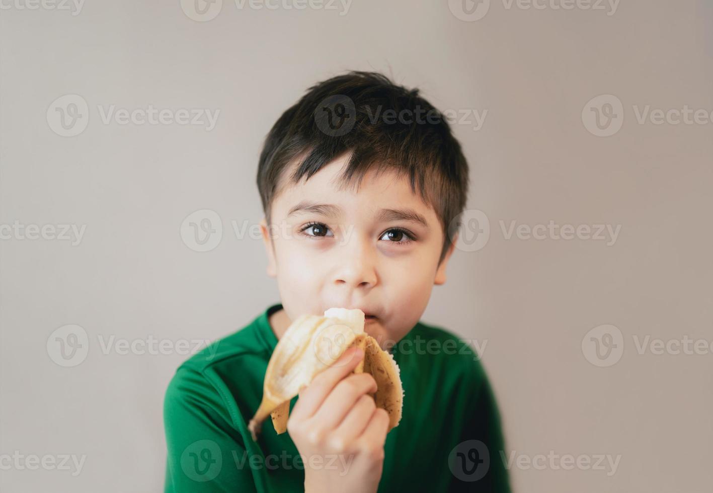 niño sano comiendo plátano, niño feliz desayunando, niño lindo mirando la cámara con cara sonriente. foto