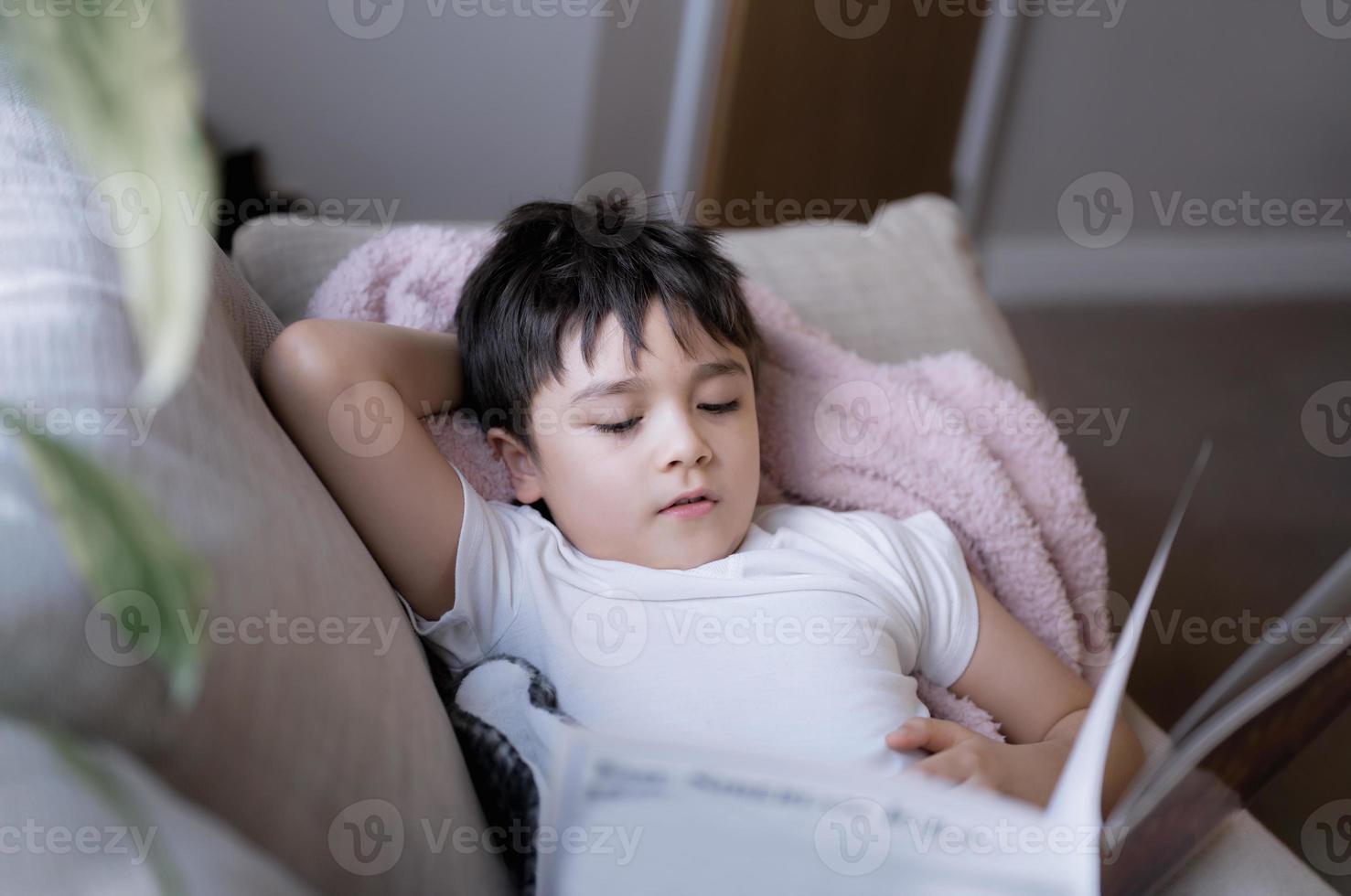Happy School kid lying on sofa reading a book with morning light shining from window. Child boy reading story relaxing at home on weekend, Positive children concept photo