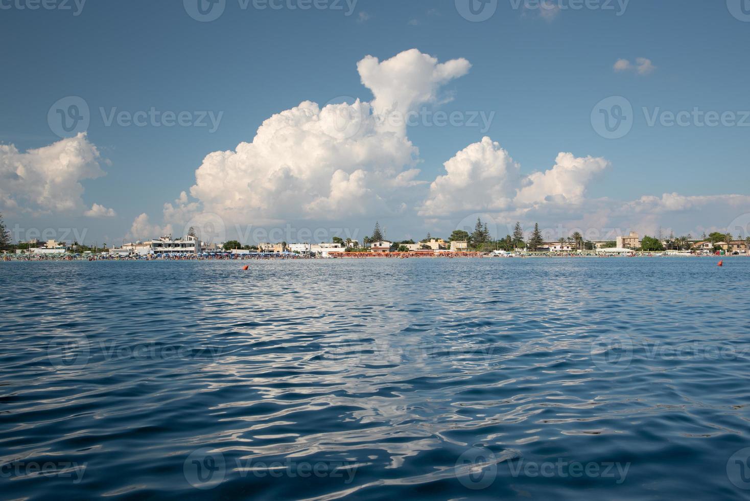 Landscape shot of the Mediterranean Sea and the water in the foreground. On the horizon a coast with the beach and many umbrellas. Above is the blue sky with white clouds reflected in the water. photo